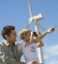 A man kneels with his son, who looks into the distance with binoculars, in front of a wind turbine. 